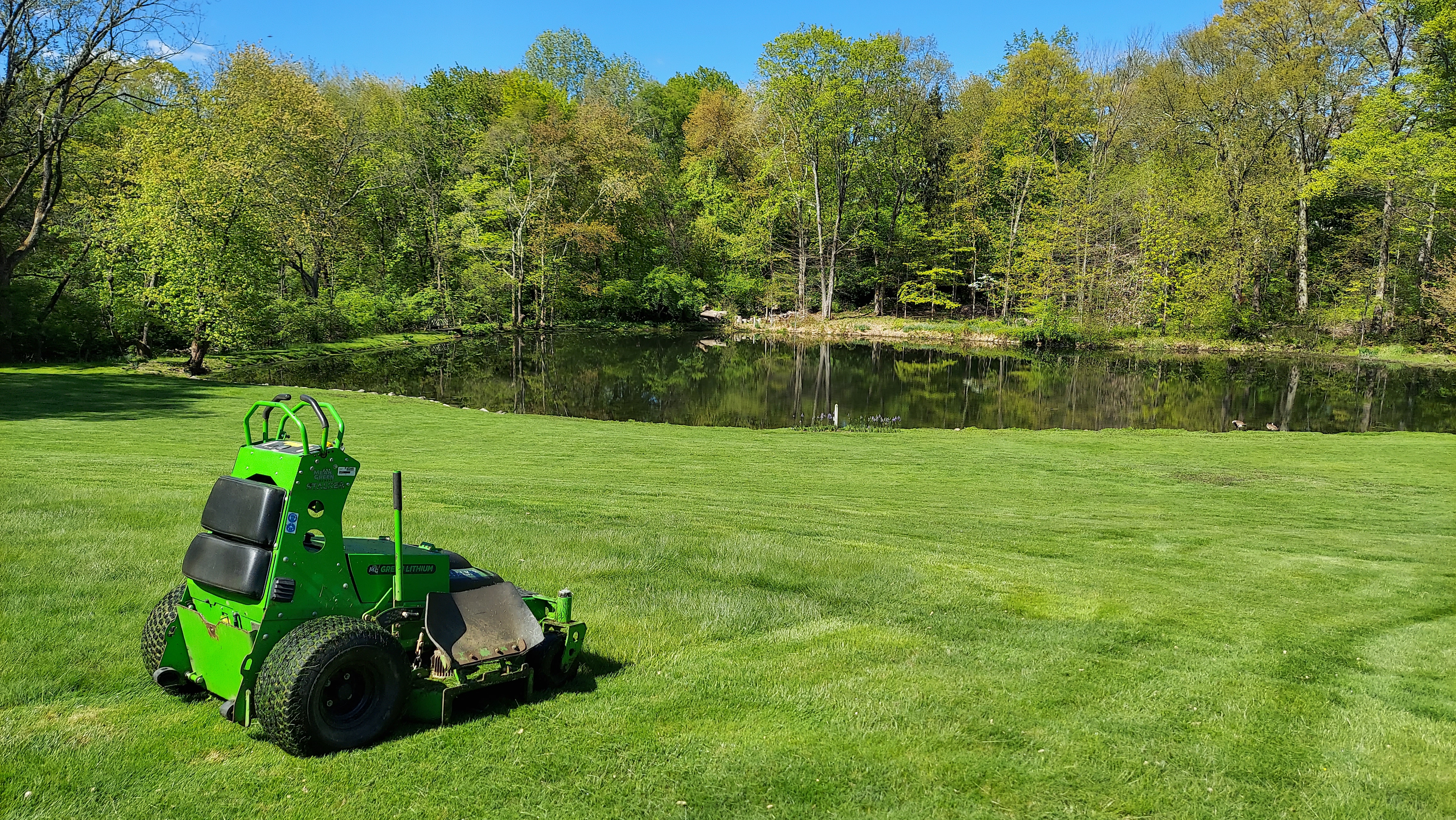 OWM electric mower on a perfectly maintained spring lawn beside a reflective pond — Fairfield County, CT